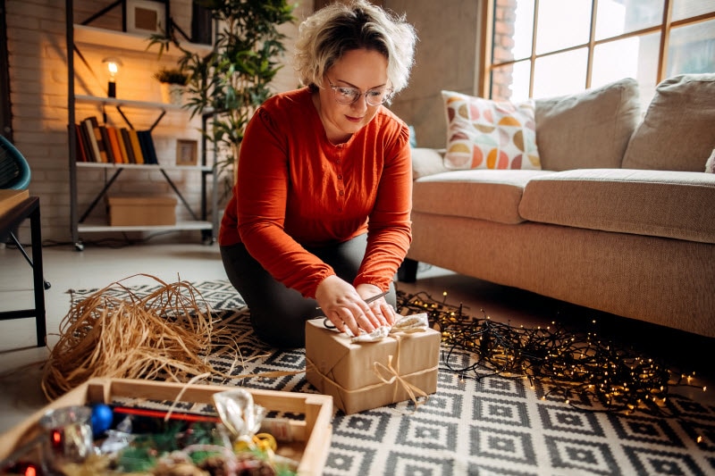 From Gift Lifting to Long Flights - What Causes Back Pain During the Holidays Woman Hunched Over on Floor Wrapping Gifts Not Understanding What Causes Holiday Back Pain