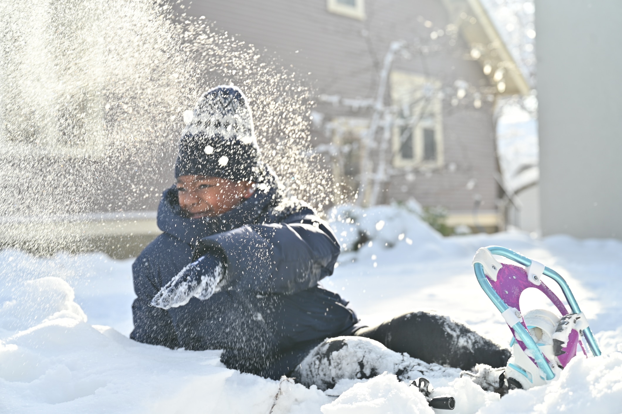Boy playing in the snow in front of a house. He has snow shoes on and the morning sun is shining behind him. He took a handful of snow and threw it on the air. The photo showed that he was smiling and having fun. The photo was taken after days of snow blizzard in Saint Paul, Minnesota.