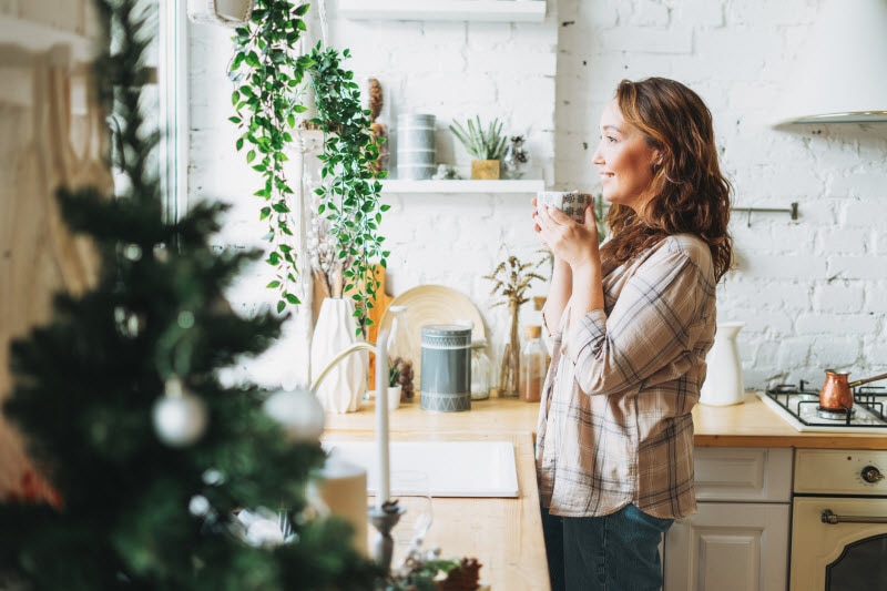 How to Manage Menopause Symptoms During the Holiday Season Woman Sipping Tea While Looking Out Kitchen Window to Represent Concept of How to Manage Menopause Symptoms During the Holidays