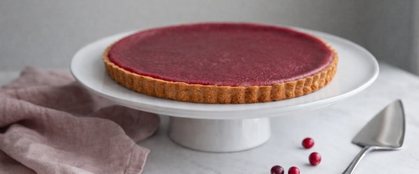 Cranberry Orange Tart with Einkorn Crust Festive Cranberry Orange Tart on White Cake Platter Set on Marble Table With Pink Napkin and Scattered Berries