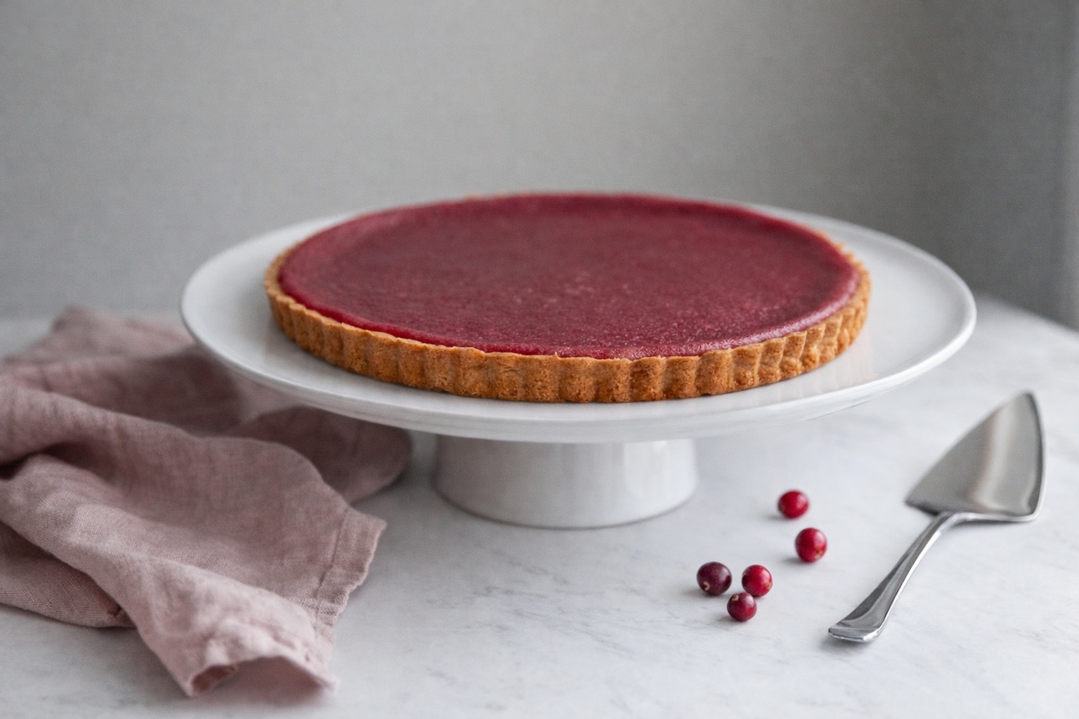 Festive Cranberry Orange Tart on White Cake Platter Set on Marble Table With Pink Napkin and Scattered Berries