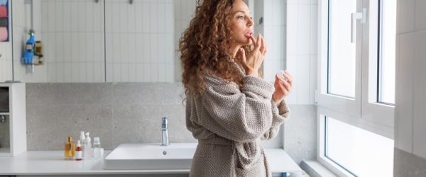 Lip Care A Woman Applies Lip Moisturizer While Looking Out the Window, Representing Lip Care During the Winter Months.