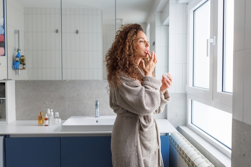 A Woman Applies Lip Moisturizer While Looking Out the Window, Representing Lip Care During the Winter Months.