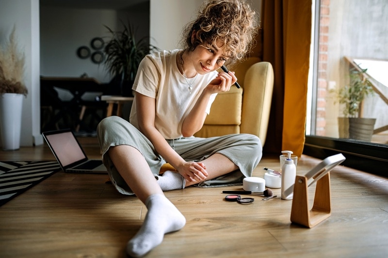 A Young Woman Applies Makeup While Sitting on Her Bedroom Floor, Representing Makeup Trends 2025.