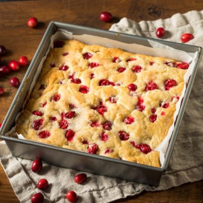A Vanilla Cake Dotted with Red Cranberries in a Metal Baking Pan Lined with Parchment Paper, all Set on a Wooden Tabletop with a Soft Linen Kitchen Towel Strewn with Spilled Fresh Cranberries (Oops!)
