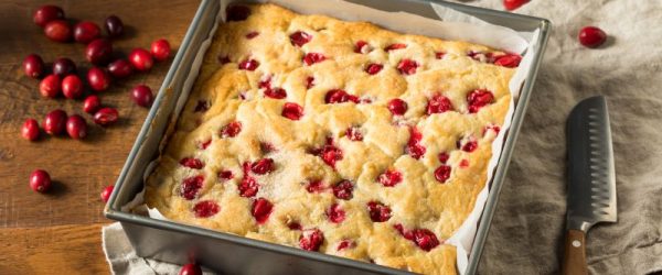 A Vanilla Cake Dotted with Red Cranberries in a Metal Baking Pan Lined with Parchment Paper, all Set on a Wooden Tabletop with a Soft Linen Kitchen Towel Strewn with Spilled Fresh Cranberries (Oops!)