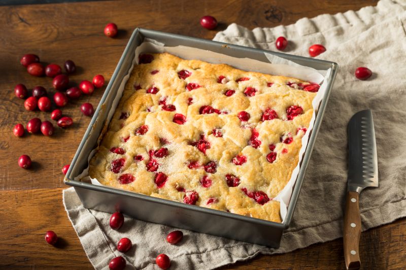 A Vanilla Cake Dotted with Red Cranberries in a Metal Baking Pan Lined with Parchment Paper, all Set on a Wooden Tabletop with a Soft Linen Kitchen Towel Strewn with Spilled Fresh Cranberries (Oops!)