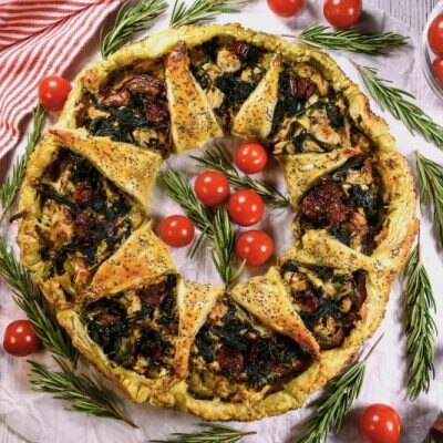 Golden Baked Puff Pastry Wreath Filled with Spinach, Artichoke Hearts, Tomato and Walnuts Laid Out on a Tabletop with a Christmas Tea Towel and Strewn Ribbons, Sprigs of Rosemary and Cherry Tomatoes Dot the Tablescape