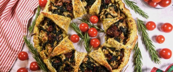 Golden Baked Puff Pastry Wreath Filled with Spinach, Artichoke Hearts, Tomato and Walnuts Laid Out on a Tabletop with a Christmas Tea Towel and Strewn Ribbons, Sprigs of Rosemary and Cherry Tomatoes Dot the Tablescape