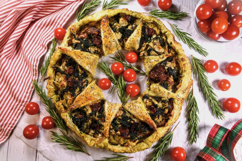 Golden Baked Puff Pastry Wreath Filled with Spinach, Artichoke Hearts, Tomato and Walnuts Laid Out on a Tabletop with a Christmas Tea Towel and Strewn Ribbons, Sprigs of Rosemary and Cherry Tomatoes Dot the Tablescape