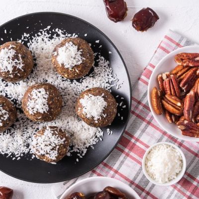 Walnut, pecan and date no-bake energy balls sprinkled with shredded coconut on a round plate, nearby are a linen tea towel and bowls of dates and pecans