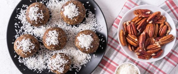 Walnut & Pecan Energy Balls_Resized Image_Liana Walnut, pecan and date no-bake energy balls sprinkled with shredded coconut on a round plate, nearby are a linen tea towel and bowls of dates and pecans