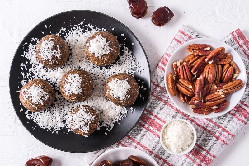 Walnut, pecan and date no-bake energy balls sprinkled with shredded coconut on a round plate, nearby are a linen tea towel and bowls of dates and pecans