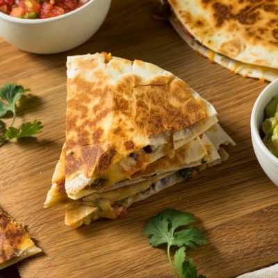 Mushroom quesadillas sliced in quarters set on a serving board surrounded by salsa, cilantro and guacamole