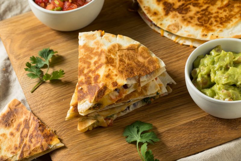 Mushroom quesadillas sliced in quarters set on a serving board surrounded by salsa, cilantro and guacamole