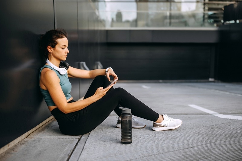 A Woman in Workout Clothes Sits and Reviews Information on Her Phone, Representing a Personalized Workout Program Created by AI.