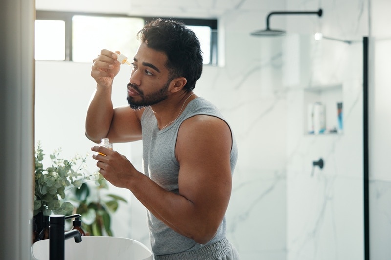 A Man Applies a Serum While Looking in the Mirror, Representing the Idea of Skin Training to Apply Active Ingredients.