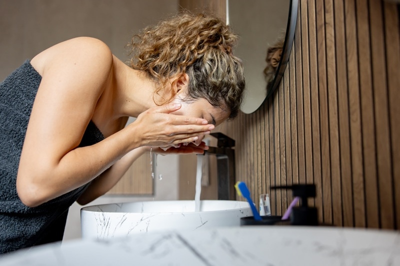 A Woman Rinses Her Face in the Sink, Representing the Effects of Hard Water on Skin.